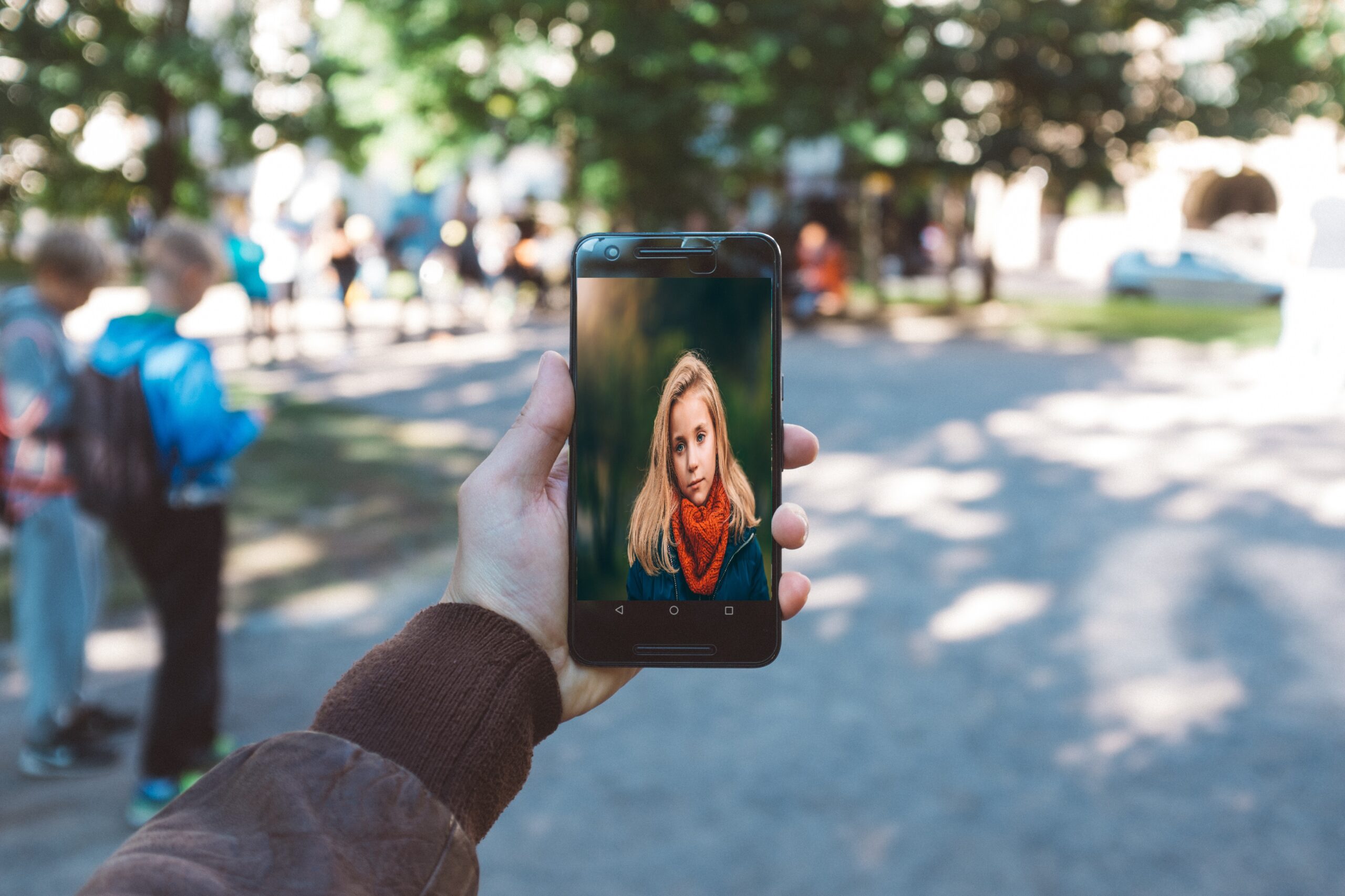 A man's arm holding a cell phone with the image of a young girl. Background shows a park with multiple children.